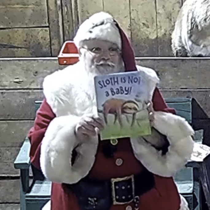 Santa standing holding book titled "Sloth is Not a Baby"